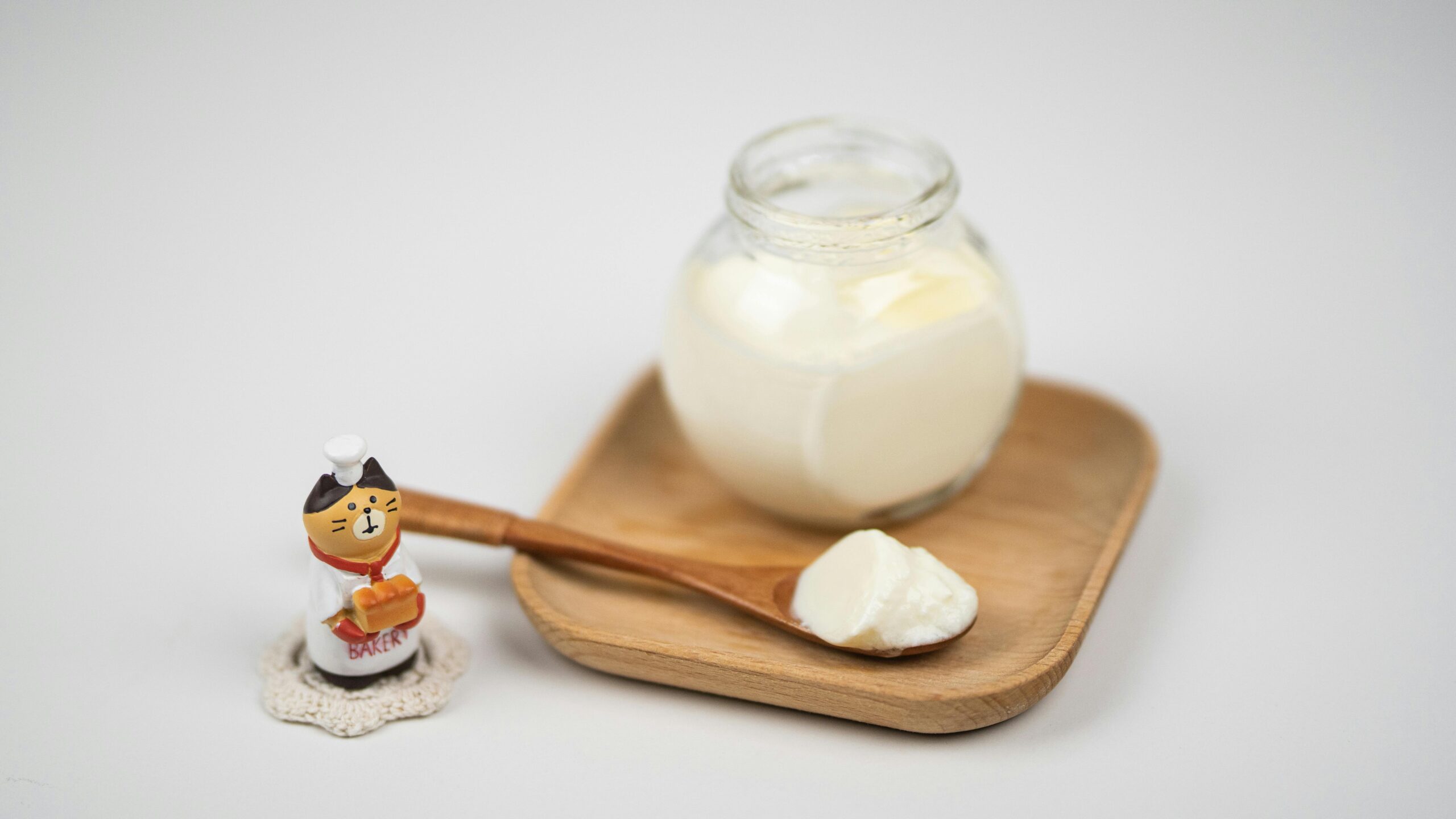 A jar of fresh yogurt on a wooden tray with a spoon, next to a cute baker figurine.